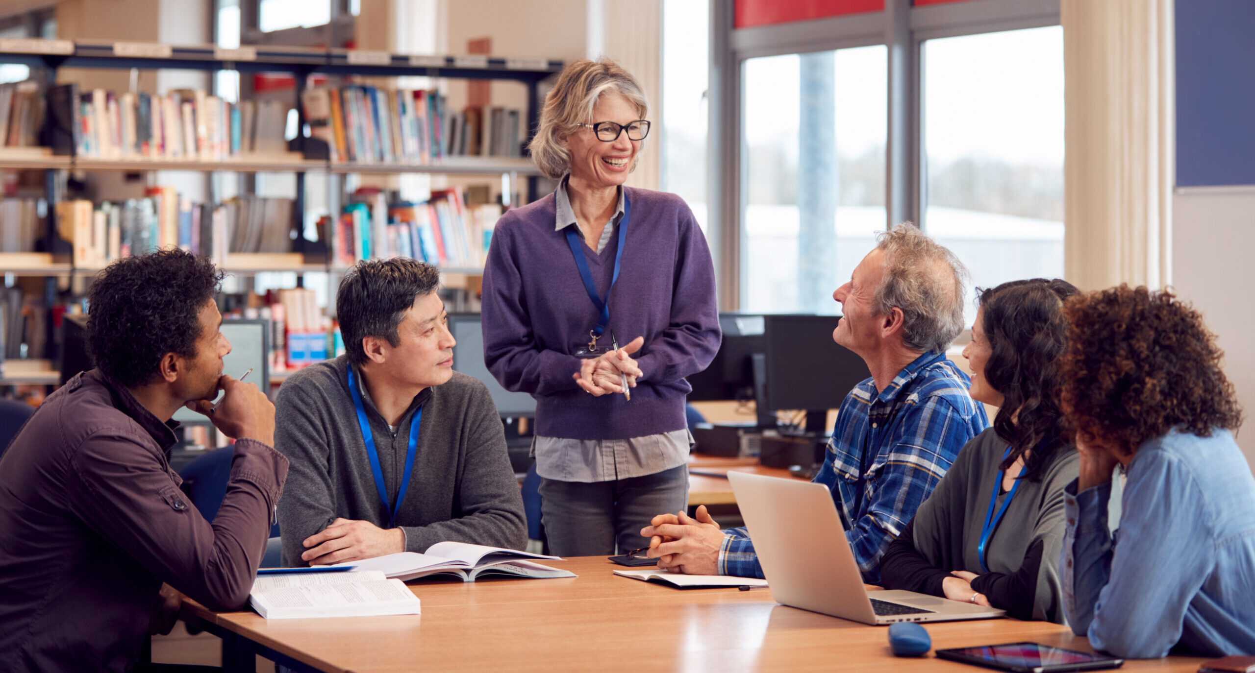 Teacher With Group Of Mature Adult Students In Class Sit Around Table And Work In College Library